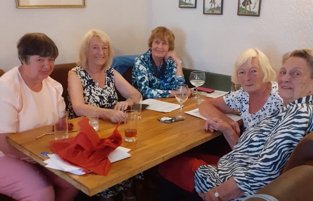 5 smiling women sitting around a table with glasses in front of them.